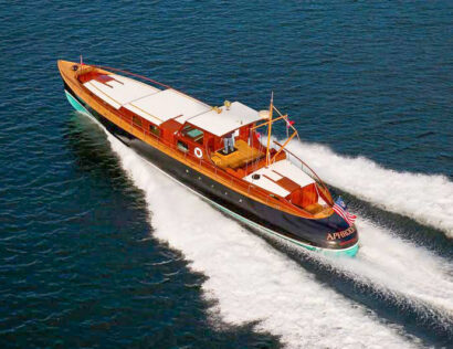 A classic wooden motor yacht cruises swiftly on blue water, leaving white waves in its wake. An American flag is visible at the stern.